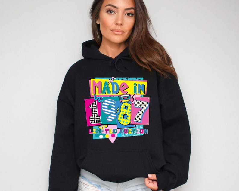 Może przedstawiać: Czarna bluza z kapturem z kolorową grafiką z napisem "MADE IN 1987 LIMITED EDITION". Grafika ma retro design z żywymi kolorami, w tym r&oacute;żowym, ż&oacute;łtym, niebieskim i zielonym. Bluza ma kaptur ze sznurkiem.