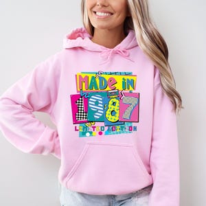 Może przedstawiać: Jasnor&oacute;żowa bluza z kapturem z kolorową grafiką z przodu z napisem "MADE IN 1987 LIMITED EDITION". Grafika ma styl retro, z cyfrą 8 w kolorze ż&oacute;łtym i turkusowym obrysem.