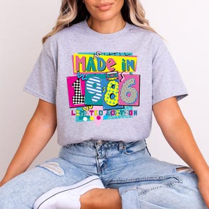 May include: A light gray t-shirt with a colorful graphic that reads "MADE IN 1986 LIMITED EDITION." The graphic features the numbers 1, 9, and 6 in different patterns and colors, including black and white checkerboard and yellow polka dots.