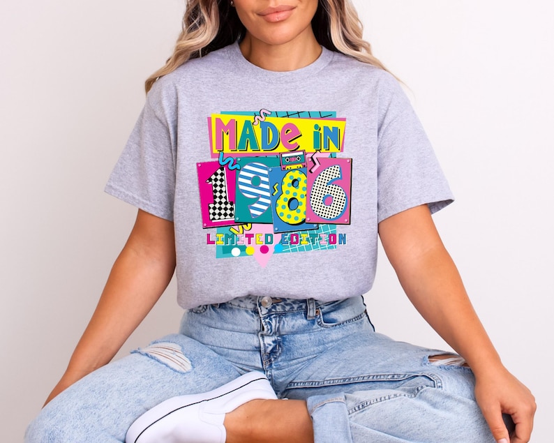 May include: A light gray t-shirt with a colorful graphic that reads "MADE IN 1986 LIMITED EDITION." The graphic features the numbers 1, 9, and 6 in different patterns and colors, including black and white checkerboard and yellow polka dots.