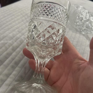 May include: Clear, vintage-style stemmed glass with diamond and textured patterns. The glass has a wide, patterned rim, a textured body, and a slender stem leading to a circular base. The glass is held in a hand.