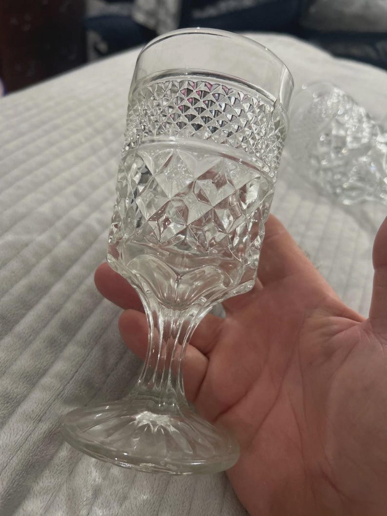 May include: Clear, vintage-style stemmed glass with diamond and textured patterns. The glass has a wide, patterned rim, a textured body, and a slender stem leading to a circular base. The glass is held in a hand.