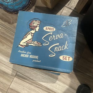 Op de afbeelding: Een vintage blauwe doos voor een "Serva-Snack" set met een illustratie van een vrouw in een gestreepte top. De doos heeft de tekst "8 Piece" en "Another fine Anchor Hocking product". De doos is licht versleten en heeft een handgeschreven nummer.