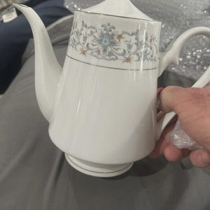 May include: White porcelain teapot with a floral design in blue, gray, and orange. The teapot has a curved spout, a handle, and a lid with a silver trim. The teapot is likely part of a tea set.
