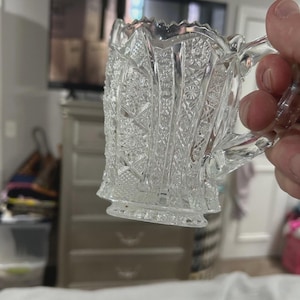 Puede incluir: Cremera de cristal tallado transparente con asa. La cremera presenta un patrón geométrico detallado y un borde festoneado. El asa también es de cristal tallado. La cremera se sostiene frente a un fondo borroso.