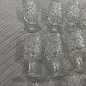 May include: A collection of clear, vintage-style water goblets. The glasses have a diamond-cut pattern on the bowl and a stemmed base. The goblets are arranged on a light gray surface.