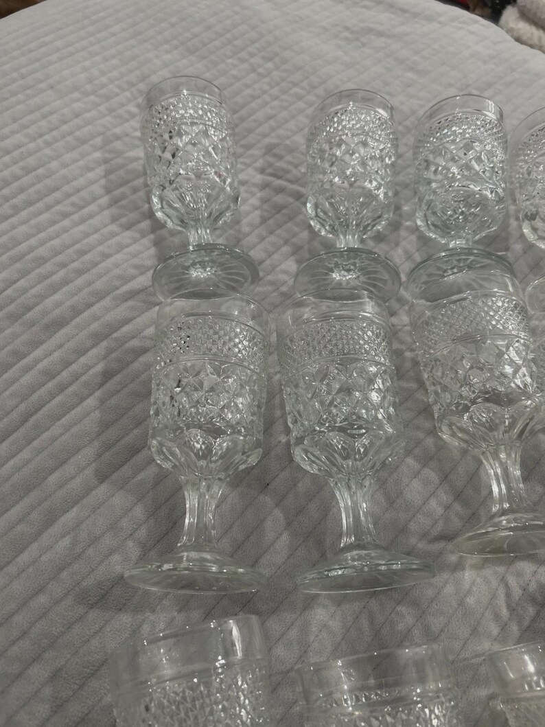 May include: A collection of clear, vintage-style water goblets. The glasses have a diamond-cut pattern on the bowl and a stemmed base. The goblets are arranged on a light gray surface.