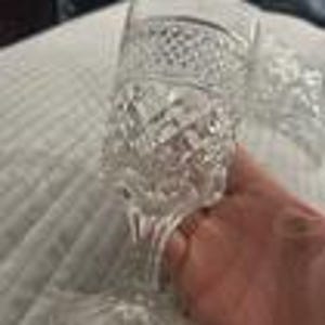 May include: A clear, cut-glass goblet with a diamond pattern and a textured stem. The goblet is held by a hand, and the background is a white, textured surface.