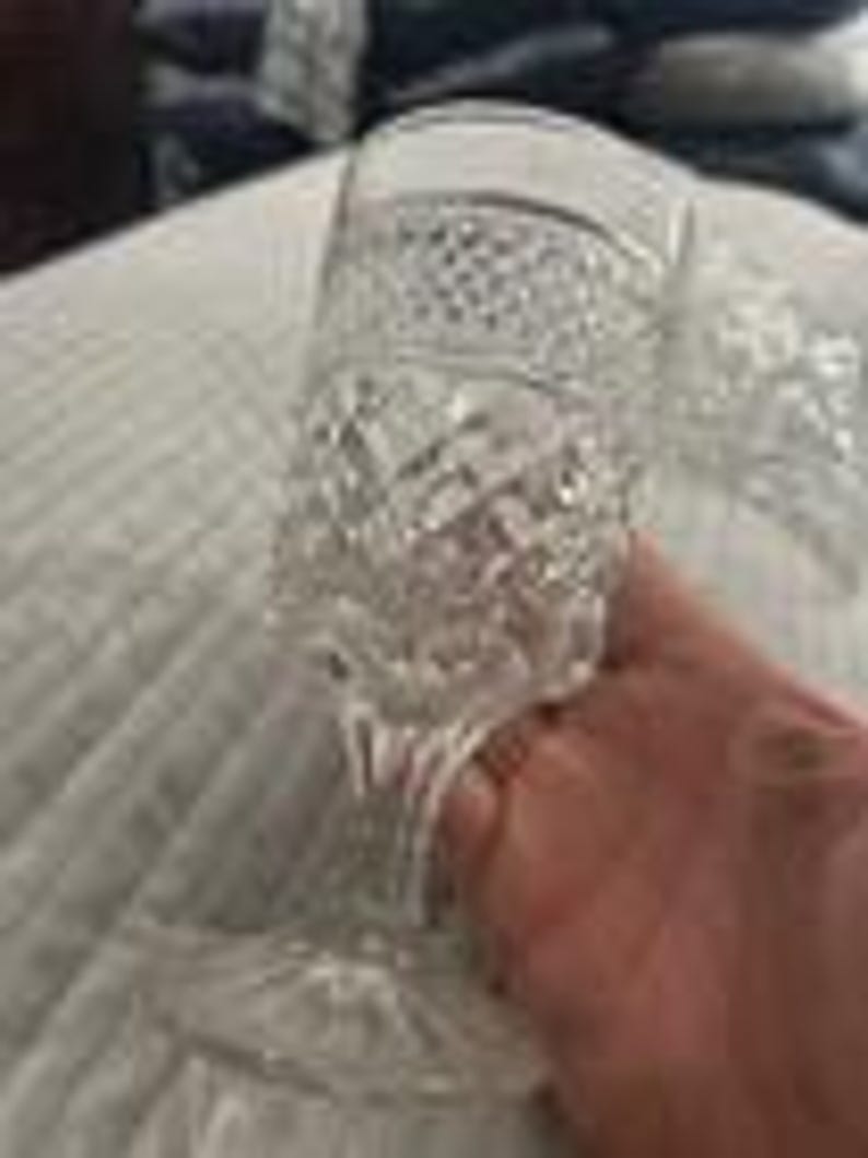 May include: A clear, cut-glass goblet with a diamond pattern and a textured stem. The goblet is held by a hand, and the background is a white, textured surface.
