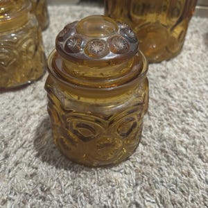 Pode incluir: Pote de vidro âmbar com tampa decorativa. O pote tem um corpo redondo com padrões em relevo e uma tampa a condizer com um design de estrela. O pote é provavelmente vintage e faz parte de um conjunto.