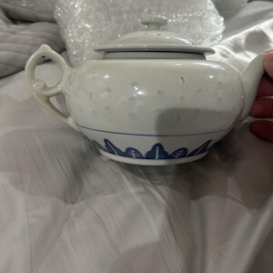 May include: A white ceramic teapot featuring a delicate embossed dot pattern on the upper body and a blue painted leaf motif around the base. The teapot includes a matching lid with a small knob handle and a curved handle with a decorative cutout.