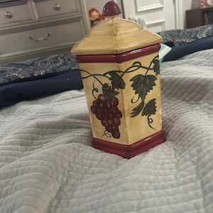 Pode incluir: Um recipiente de cerâmica quadrado com corpo na cor creme e base e tampa em vermelho escuro. O recipiente apresenta um design pintado de uvas roxas e folhas de videira verdes na parte frontal. Possui um pequeno puxador arredondado no topo da tampa.