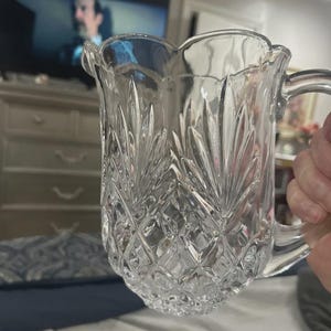 May include: Clear glass pitcher with a decorative, cut-glass design. The pitcher has a handle and a scalloped rim. The cut-glass pattern features diamond shapes and leaf-like designs. The pitcher is held by a person.