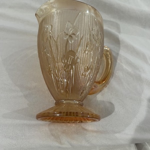 May include: A vintage amber glass pitcher with a floral design. The pitcher has a flared base, a handle, and a spout. The glass has a textured surface with vertical lines and embossed flowers. The pitcher is likely used for serving beverages.