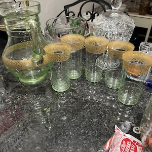 May include: A collection of vintage glassware, including a green glass pitcher with gold trim, several clear drinking glasses with gold rims, and a crystal candy dish. The glassware is arranged on a dark countertop.