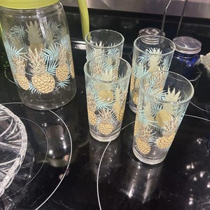 May include: A clear glass pitcher and matching drinking glasses with a pineapple and palm leaf design. The pitcher has a green lid and handle. The glasses and pitcher are decorated with gold pineapples and blue and green leaves.