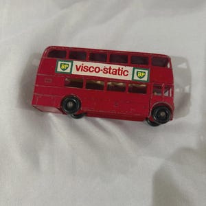 May include: A small, red double-decker bus toy with black wheels. The bus features the text "visco-static" in white lettering, flanked by green and white "BP" logos. The toy is a classic design, likely a collectible item.