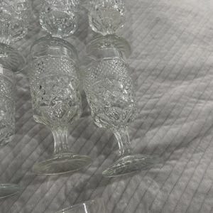 May include: A collection of clear, vintage-style glassware. The glasses feature a diamond-cut pattern and are of varying shapes, including stemmed wine glasses and tumblers. The glassware is arranged on a light gray surface.