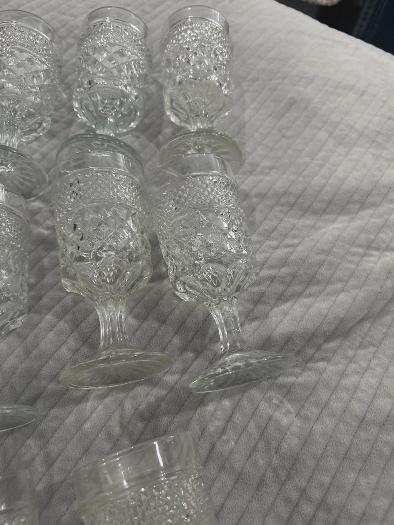 May include: A collection of clear, vintage-style glassware. The glasses feature a diamond-cut pattern and are of varying shapes, including stemmed wine glasses and tumblers. The glassware is arranged on a light gray surface.