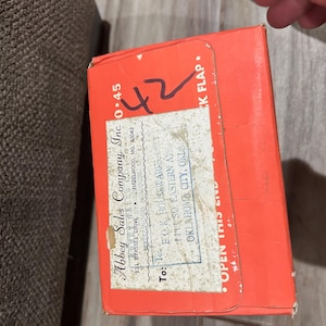 Op de afbeelding: Een feloranje doos met de tekst "Abbey Sales Company, Inc." en handgeschreven cijfers. De doos heeft een verzendlabel met een adres en de tekst "OPEN THIS END".