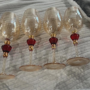 Könnte beinhalten: Vier elegante Weingläser mit bernsteinfarbenen Kelchen und klaren Stielen. Jedes Glas hat einen roten, bauchigen Fuß mit goldenen Akzenten. Die Kelche weisen ein dezentes Sternmuster auf, das dem Glaswaren einen Hauch von Raffinesse verleiht.