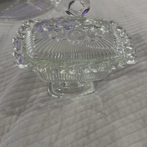May include: A clear glass candy dish with a lid. The dish is square with a scalloped edge and a ribbed design. The lid has a decorative knob. The dish sits on a pedestal base. The overall design is vintage.