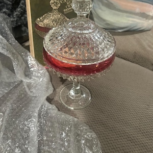 Peut inclure: Un bonbonnier en verre transparent et rouge rubis avec un couvercle et un bol à motif diamant, reposant sur un pied. Le couvercle présente un fleuron pointu. La boîte d'origine est visible en arrière-plan, marquée Diamond Point Candy Ruby Crystal.