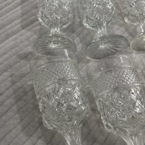 May include: A collection of clear, stemmed glassware with diamond-cut patterns. The glasses have a classic design, featuring a rounded bowl and a slender stem. The glassware is arranged on a soft, textured surface.