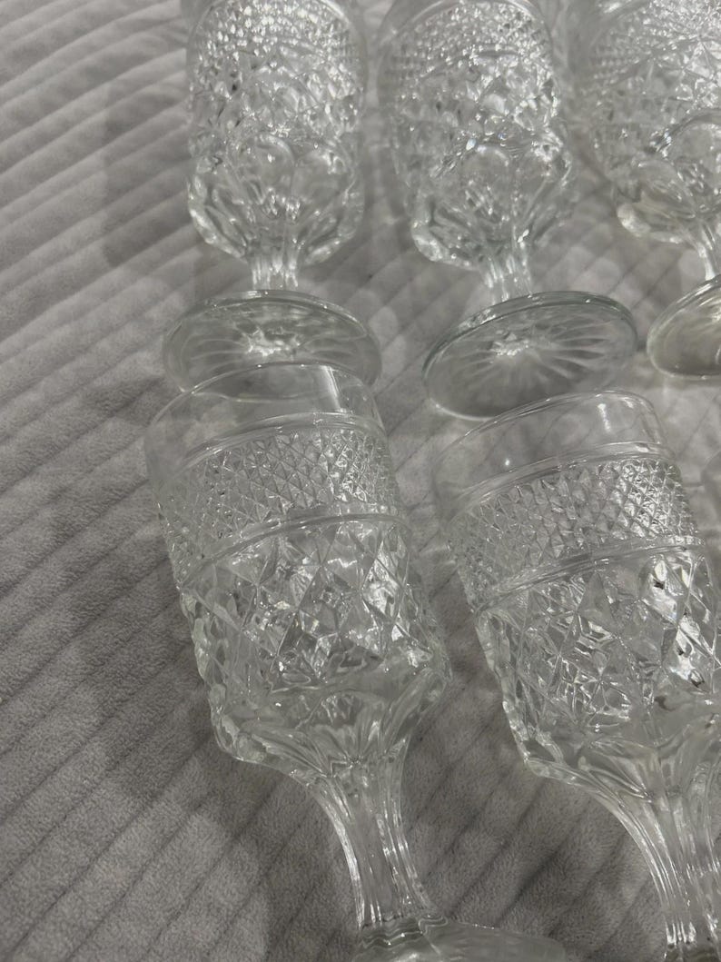 May include: A collection of clear, stemmed glassware with diamond-cut patterns. The glasses have a classic design, featuring a rounded bowl and a slender stem. The glassware is arranged on a soft, textured surface.