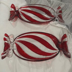 Könnte beinhalten: Zwei klare, bonbonförmige Glasschalen mit rot-weiß gestreiften Designs. Die Schalen haben an jedem Ende ein schleifenartiges Detail. Die Schalen befinden sich auf einer weißen Oberfläche.
