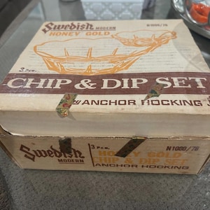 May include: A vintage "Swedish Modern Honey Gold" chip and dip set box. The box is beige with orange text and graphics, including the words "CHIP & DIP SET" and "ANCHOR HOCKING". The box is sealed with tape.