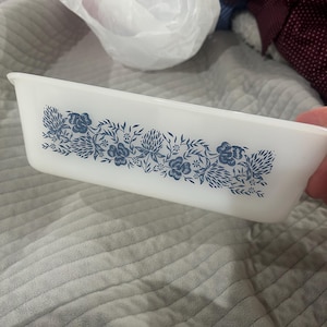 May include: A rectangular white milk glass baking dish featuring a repeating blue floral pattern along the side. The design consists of detailed illustrations of various flowers and leaves arranged in a horizontal band across the centre.