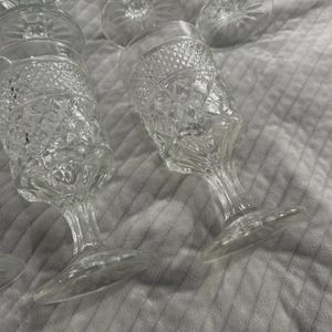 May include: Clear, stemmed glassware with diamond-patterned detailing. The glasses have a wide base and a slender stem, with a rounded bowl. Several glasses are visible, some upright and some tilted, on a textured, light-colored surface.