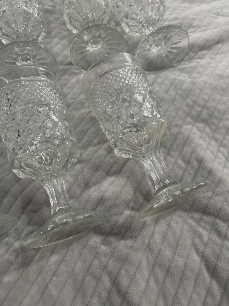 May include: Clear, stemmed glassware with diamond-patterned detailing. The glasses have a wide base and a slender stem, with a rounded bowl. Several glasses are visible, some upright and some tilted, on a textured, light-colored surface.
