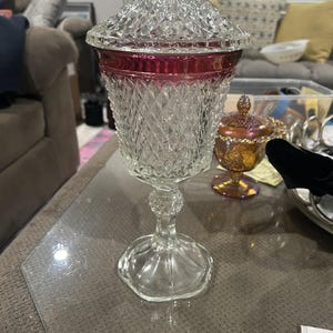 Puede incluir: Un bombonera de cristal transparente con un patrón de diamantes y una banda roja rubí alrededor de la parte superior. El plato tiene tapa y se asienta sobre una base de pedestal. Un plato de cristal iridiscente más pequeño está en el fondo.