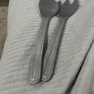 May include: A pair of silver salad servers. One is spoon-shaped, the other fork-shaped. Both have long handles with a beaded design. The servers are resting on a grey, textured surface.