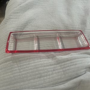 May include: A rectangular clear glass serving tray featuring three distinct compartments separated by glass dividers. The outer rim of the tray is accented with a vibrant red border that frames the entire structure of the serving dish.