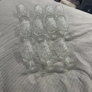 May include: A collection of twelve clear, stemmed glasses with a textured, diamond-like pattern. The glasses are arranged on a white, quilted surface. The stemware has a classic, vintage aesthetic.