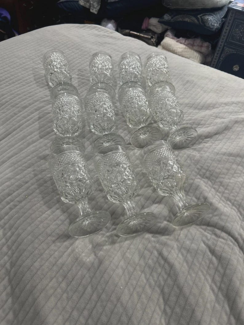 May include: A collection of twelve clear, stemmed glasses with a textured, diamond-like pattern. The glasses are arranged on a white, quilted surface. The stemware has a classic, vintage aesthetic.