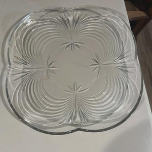 Puede incluir: Una fuente de vidrio transparente con forma de cuatro lóbulos. La fuente presenta un diseño central de líneas radiales y patrones de estrellas, creando una apariencia decorativa y elegante. La fuente está diseñada para servir alimentos.