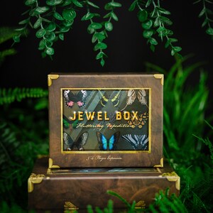 May include: A brown wooden box with a gold trim and a clear plastic window showing colorful butterfly illustrations. The box says "Jewel Box:  Fluttering Expedition" and "5-6 Player Expansion".