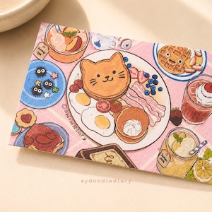 May include: A rectangular postcard featuring a colorful illustration of a breakfast spread. The central plate displays a cat-shaped pancake, fried eggs, bacon, and berries. Surrounding items include iced tea, waffles, toast with strawberries, and lemonade.
