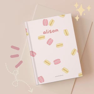 Könnte beinhalten: Ein Notizbuch mit hellrosa Einband, verziert mit rosa und gelben Makronen. Der Name "alison" ist in Rosa auf dem Einband geschrieben. Das Notizbuch liegt auf einer hellrosa und beigefarbenen Oberfläche.