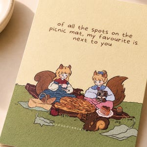 Könnte beinhalten: Eine cremefarbene Grußkarte mit einer Illustration von zwei Charakteren mit Eichhörnchenohren bei einem Picknick auf einer grünen Matte. Der Text lautet: of all the spots on the picnic mat, my favourite is next to you. Ein Korb, Brot und ein kleines Eichhörnchen sind abgebildet.