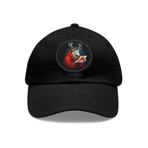 May include: Black baseball cap featuring a circular patch with a deer-like creature in a red robe reading a card. The deer has white fur and antlers. The patch is outlined with a white dashed line. The cap has a curved brim and a fabric strap closure.