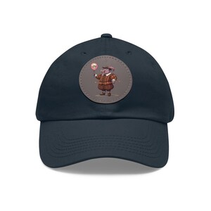 May include: Navy blue baseball cap with a circular patch featuring a cartoon pig in Renaissance attire holding a skull on a stick. The pig is wearing a brown and red outfit. The cap has a curved brim.