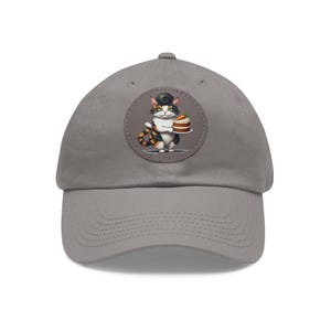 May include: A gray baseball cap with a round patch featuring a cartoon cat holding a carrot cake. The cat has black, white, and orange fur and a black pompadour.