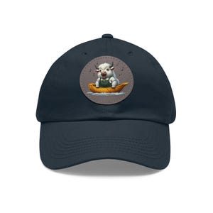 May include: A navy blue baseball cap with a circular patch featuring a white cow wearing overalls and singing in a yellow boat. The patch has a gray background and musical notes around the cow.