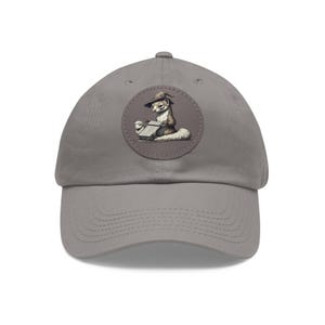 May include: A gray baseball cap with a curved brim. The cap features a circular patch with an illustration of a ferret wearing a witch hat and reading a book. The ferret is brown, white, and black.