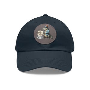 May include: Navy blue baseball cap with a round patch featuring an alligator dressed in a blue blazer and hat, packing a suitcase. The alligator is depicted in a cartoon style. The cap has a curved brim and an adjustable strap.